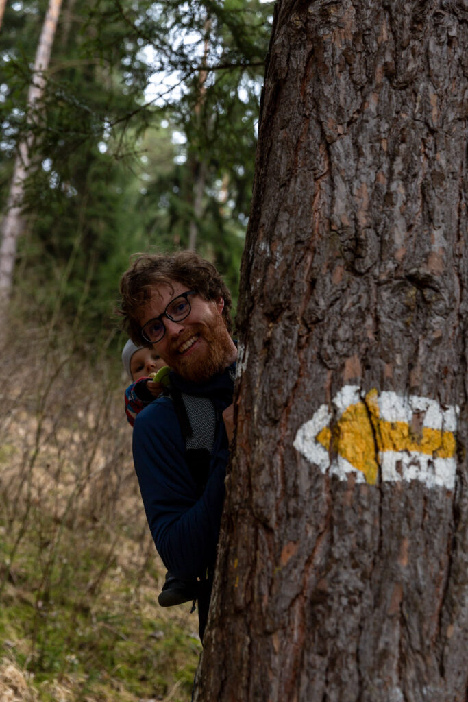 Michal a Markus v Thule Sapling detský turistický nosič. Pri strome a turistickej značke v tvare šípky na strome. Michal sa skrýva za stromom. 