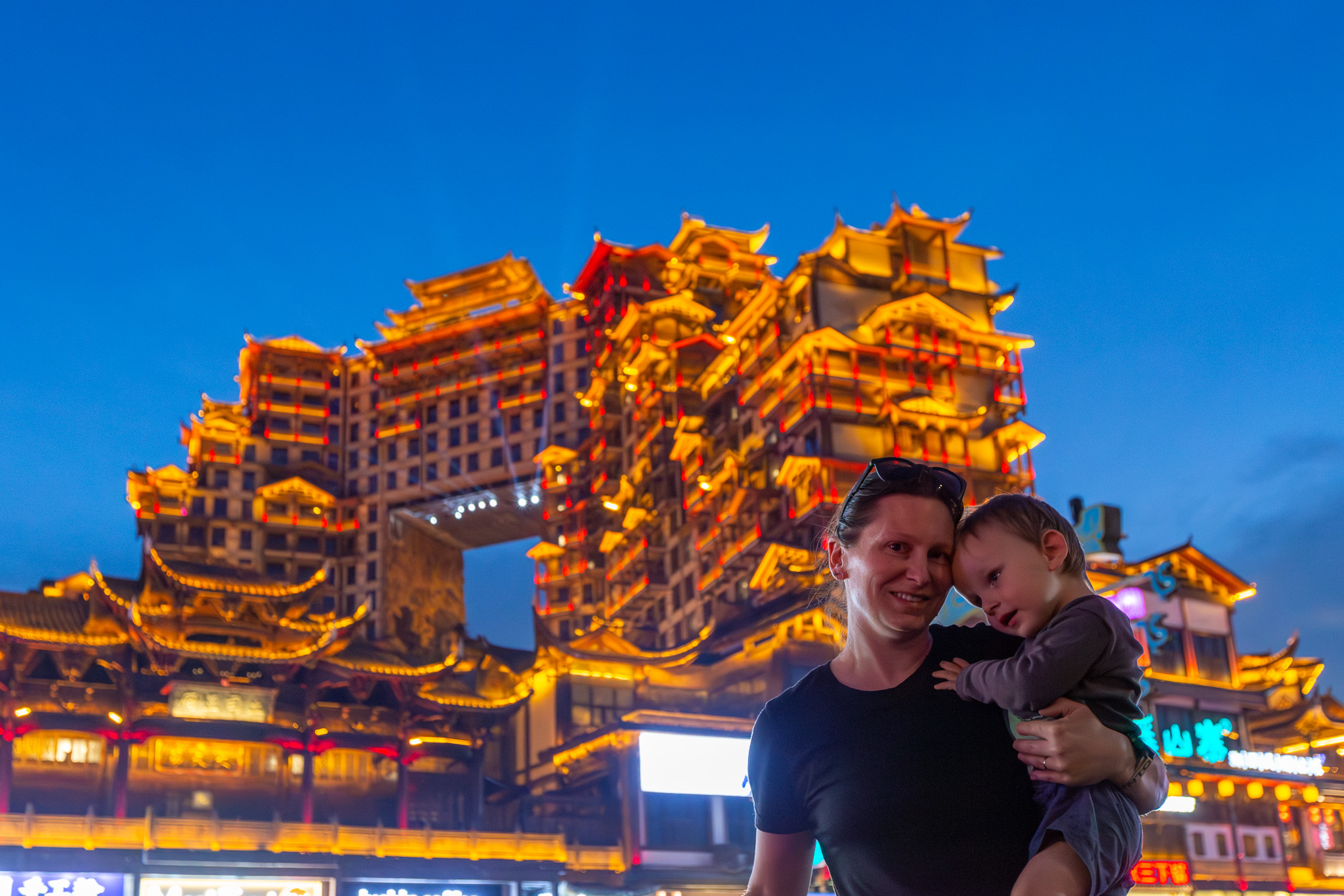 Žena držiaca dieťa pred vysvietenou budovou 72 Wonder Tower v Zhangjiajie
