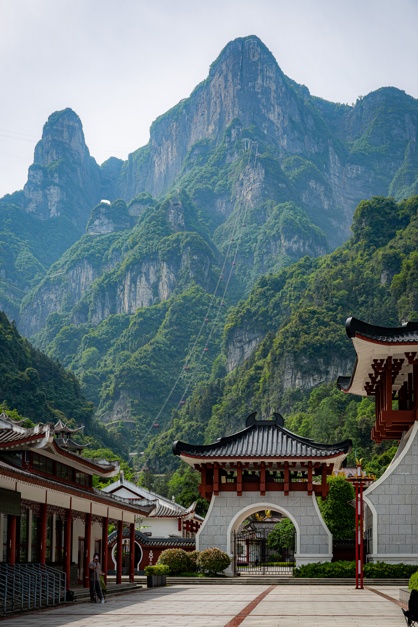 Tradičná čínska brána a architektúra v popredí, za ktorou sa týči mohutný skalný masív Tianmen Mountain s lanovkou a ikonickým oknom Nebeská brána v meste Zhangjiajie, Čína.