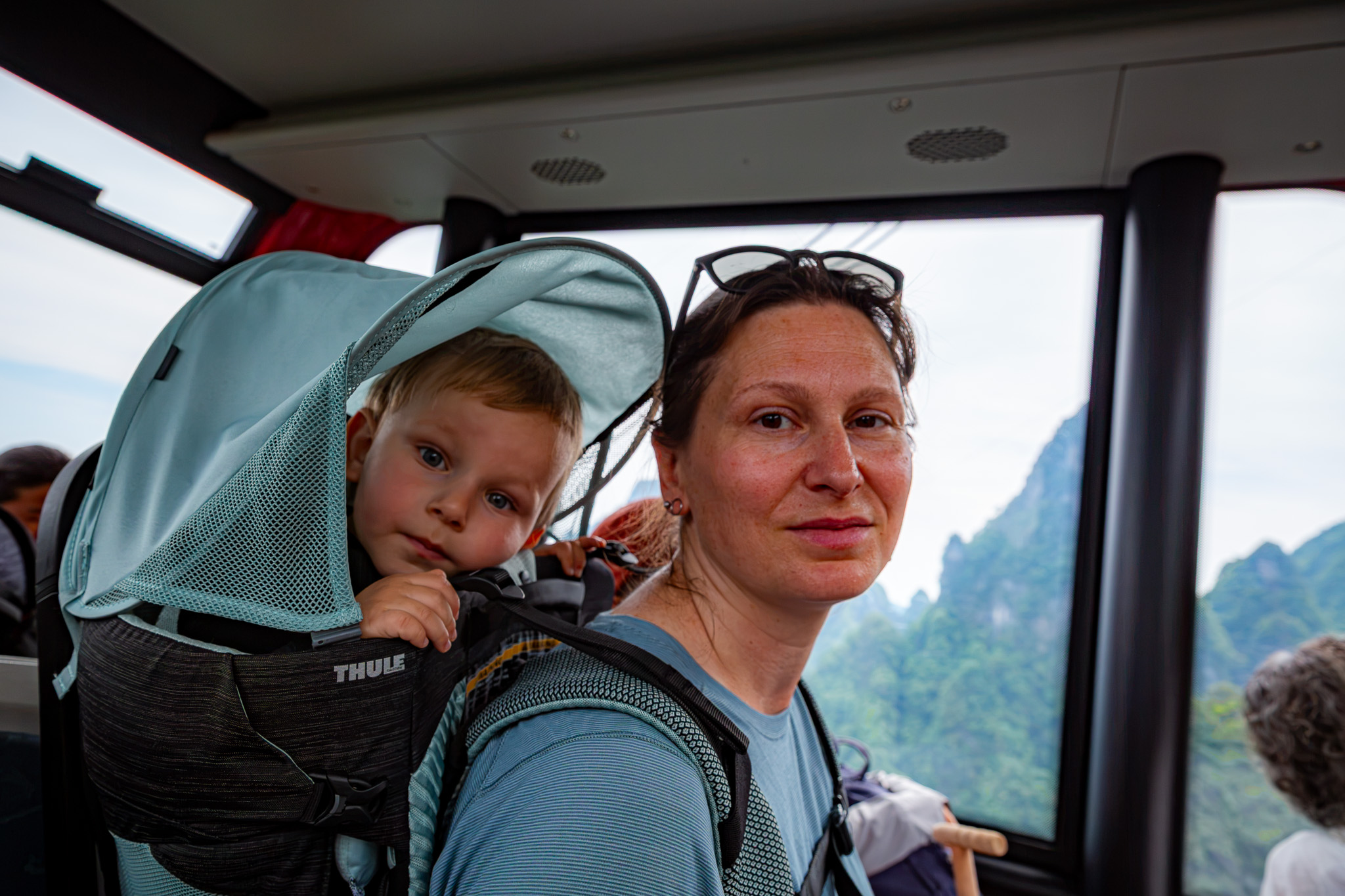 Maca so synom Markusom (matka s malým dieťaťom v turistickom nosiči) sa vezú v presklenej lanovke smerom hore na Tianmen Mountain. Cez okno v pozadí je vidieť majestátne krasové vrchy v oblasti Zhangjiajie, Čína.