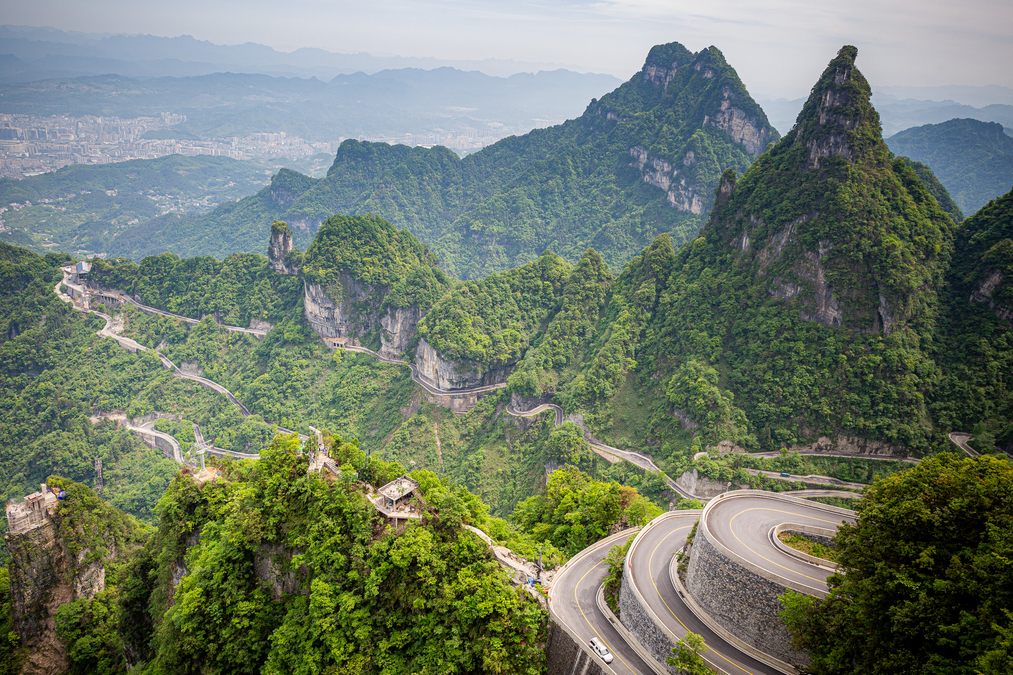 Dychberúci výhľad na majestátne krasové vrcholy a slávnu kľukatú horskú cestu s 99 zákrutami v oblasti Tianmen Mountain. V diaľke sa rozprestiera mesto Zhangjiajie, Čína.