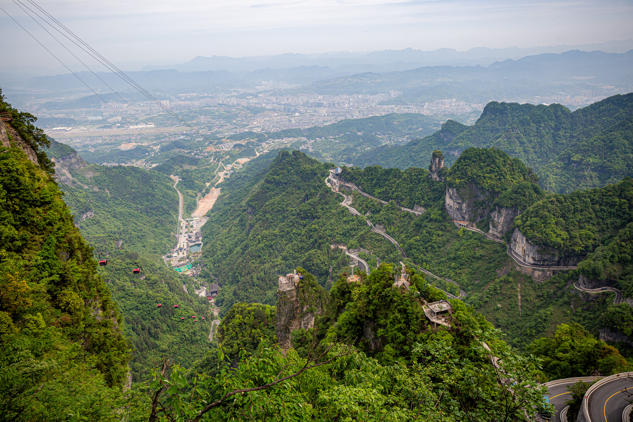 Dychberúci panoramatický výhľad z masívu Tianmen Mountain do hlbokého zeleného údolia. Na úbočí hory je vidieť slávnu kľukatú cestu, červené kabínky lanovky a v pozadí rozľahlé mesto Zhangjiajie, Čína.