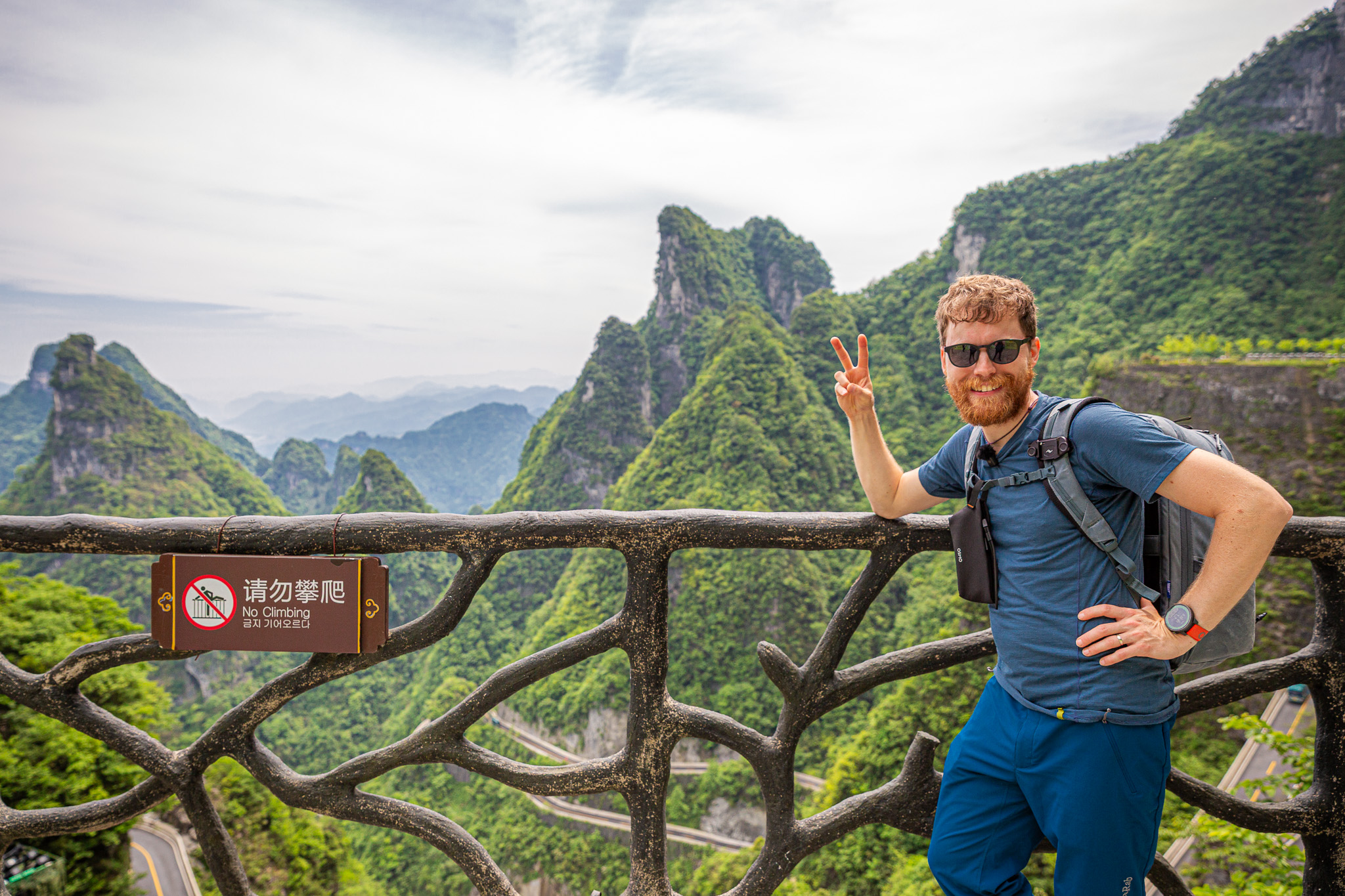Mišo (turista s batohom) sa usmieva a ukazuje znak mieru na vyhliadke. V pozadí sa týčia majestátne zelené vrchy a slávna kľukatá horská cesta pod masívom Tianmen Mountain v oblasti Zhangjiajie, Čína.