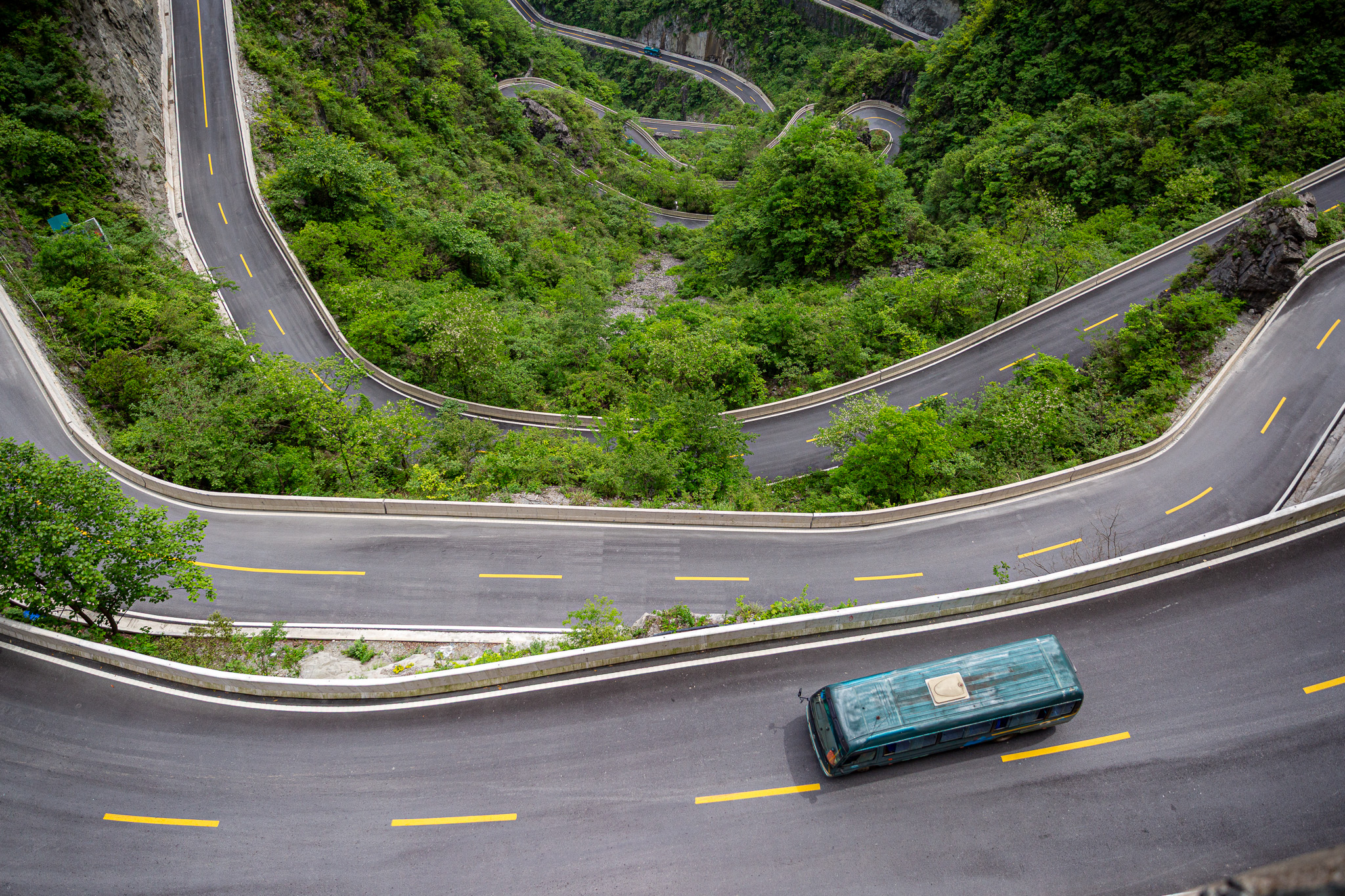 Pohľad zhora na extrémne kľukatú horskú cestu plnú ostrých zákrut, ktorá sa zarezáva do zeleného masívu Tianmen Mountain. Po asfalte prechádza turistický autobus v oblasti Zhangjiajie, Čína.