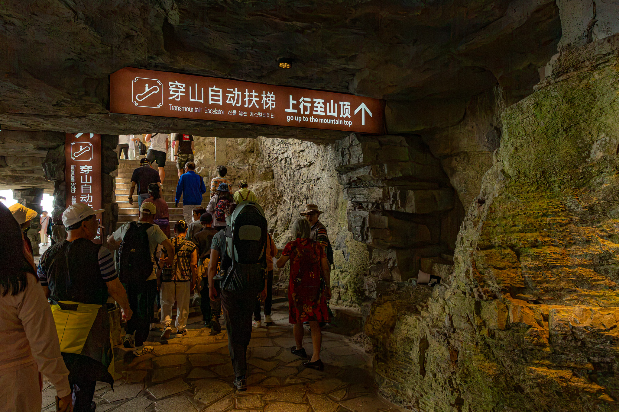 Maca so synom Markusom v turistickom nosiči (matka s malým dieťaťom) kráčajú v podzemnom skalnom tuneli k eskalátorom vedúcim na vrchol Tianmen Mountain v oblasti Zhangjiajie, Čína.