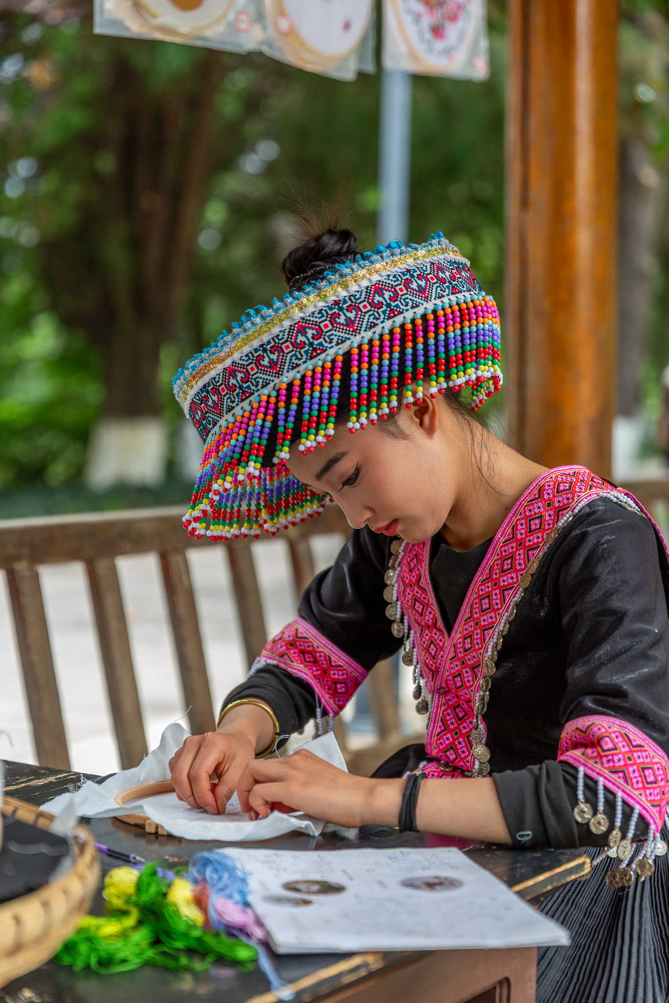 Mladá žena v tradičnom čiernom oblečení a farebnom klobúku s korálkami vyšíva za stolom, Yunnan Ethnic Village, Kunming, Čína