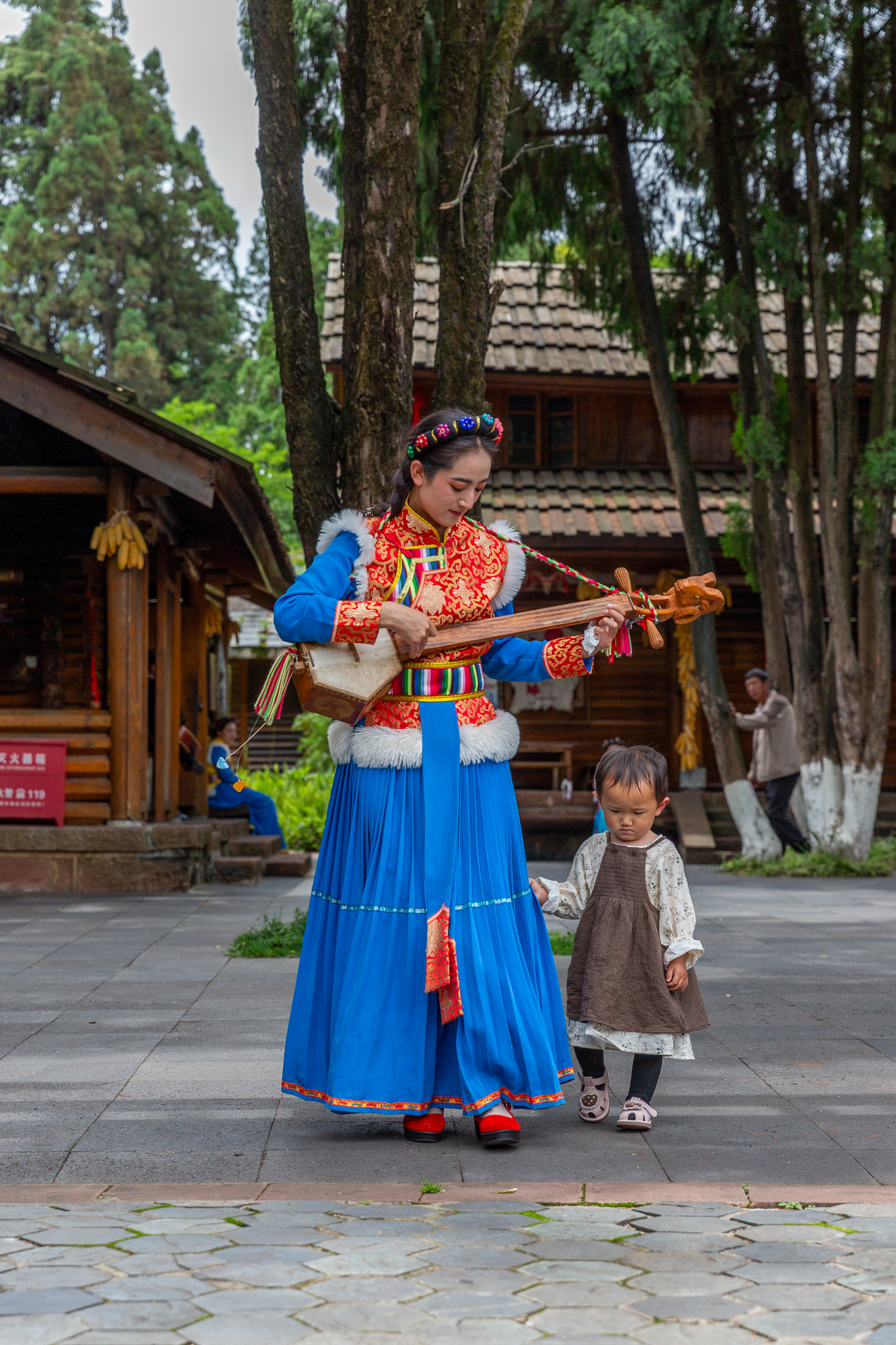 Mladá žena v tradičnom modrom oblečení hrá na hudobnom nástroji pripomínajúcom gitaru zatiaľ čo ju za sukňu drží jej malé dieťa, Yunnan Ethnic Village, Kunming, Čína