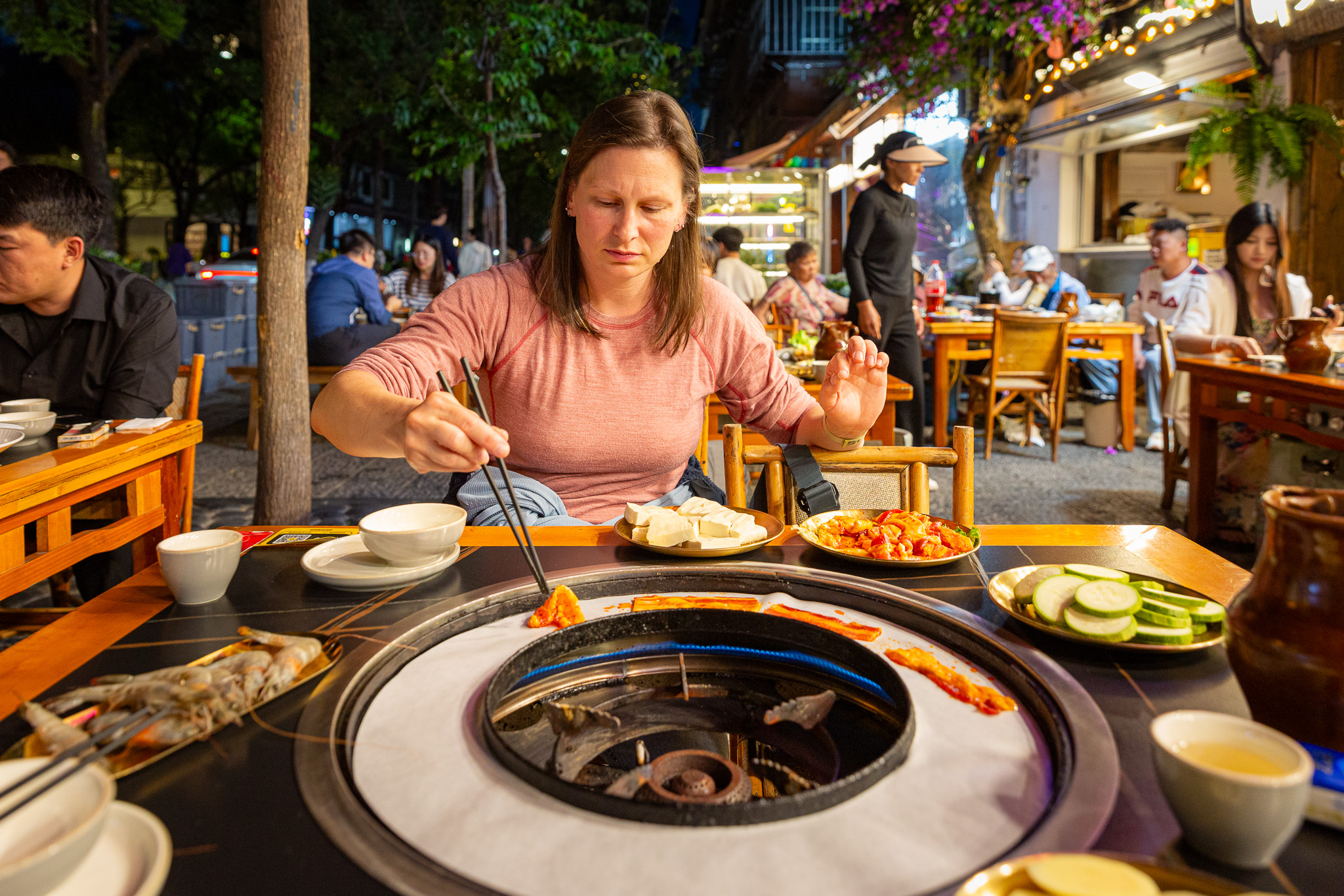 Kulinársky zážitok v provincii Yunnan, konkrétne papierové grilovanie (Paper BBQ / Zhi Shang Kao Rou) priamo v nočných uličkách historického mesta Dali. Žena nakladá suroviny na papier. Priamo na papieri sa opekajú čerstvé krevety, tenké plátky marinovaného bravčového bôčika a kolieska cukety.