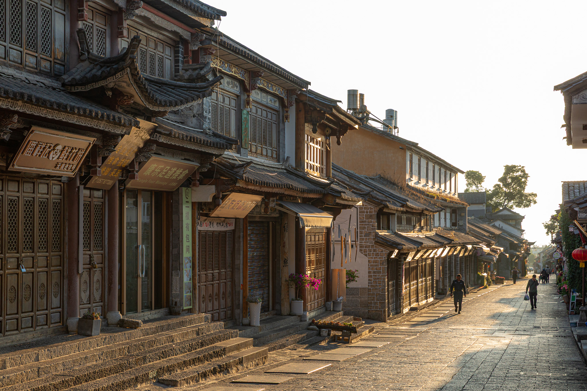 Pokojné ranné prebúdzanie v uličkách historického mesta Dali (Dali Ancient Town), kedy ranné slnko vytvára dlhé tiene na kamennom dláždení. Detailný pohľad na tradičné viacposchodové budovy s vyrezávanými drevenými dverami, okenicami a typickými zakrivenými rohmi striech. Ulica je takmer prázdna, vidno len zopár miestnych obyvateľov kráčajúcich do práce či na nákup, čo ostro kontrastuje s rušnou atmosférou, ktorá tu vládne popoludní.