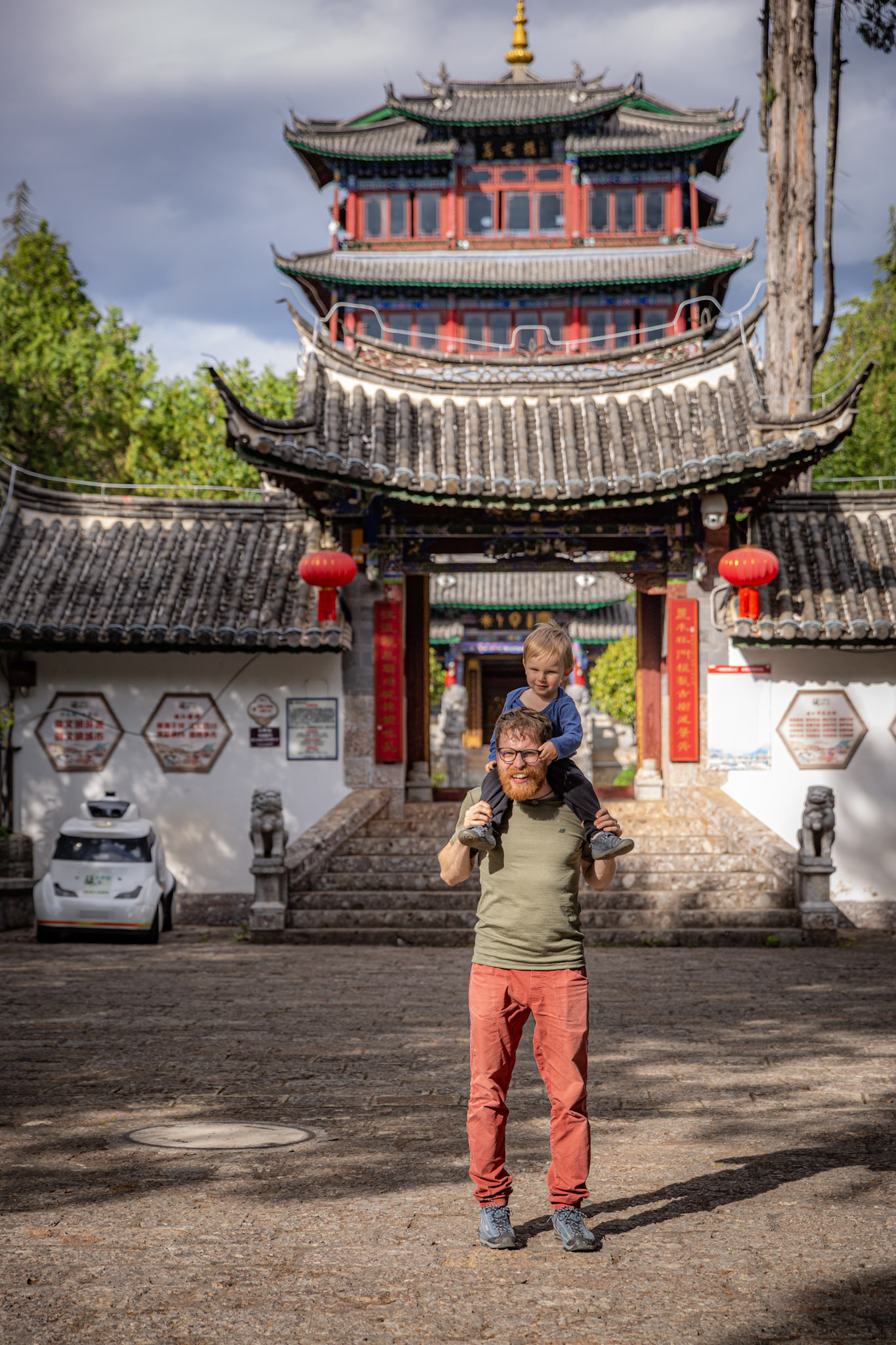 Muž v zelenom tričku s malým dieťaťom na ramenách stojí pred vežou na Shizi Mountain Wangulou, Lijiang