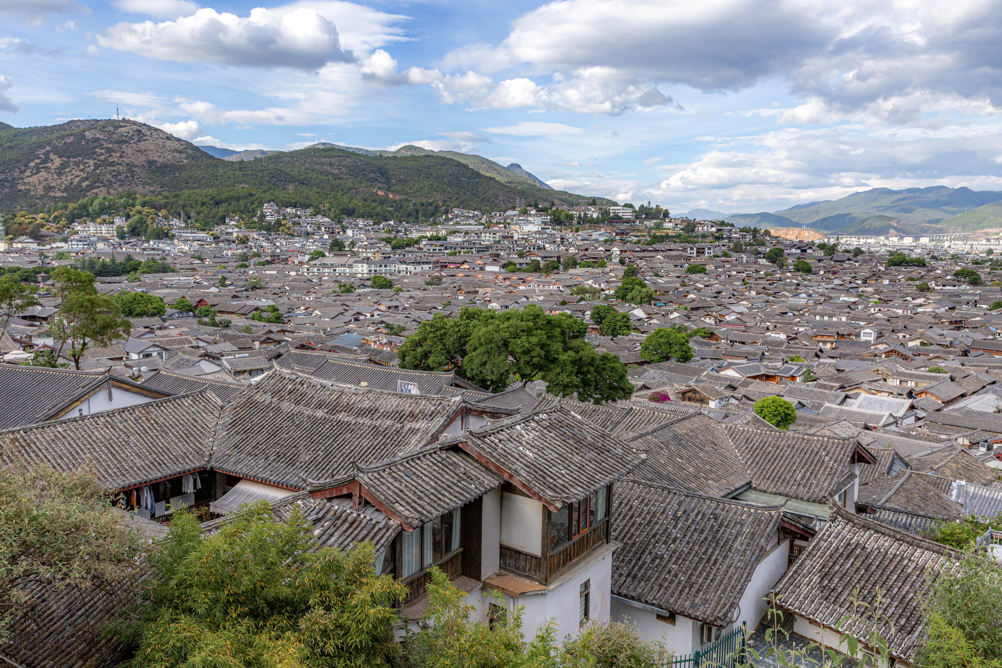 Panoráma historického mesta Lijiang s tradičnými strechami domov pod zvlnenými zelenými kopcami.