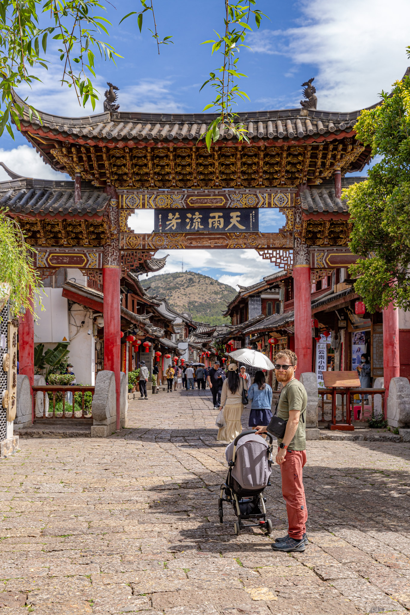 Muž s detským kočíkom stojí na kamennej uličke pred monumentálnou tradičnou čínskou bránou v meste Lijiang. 