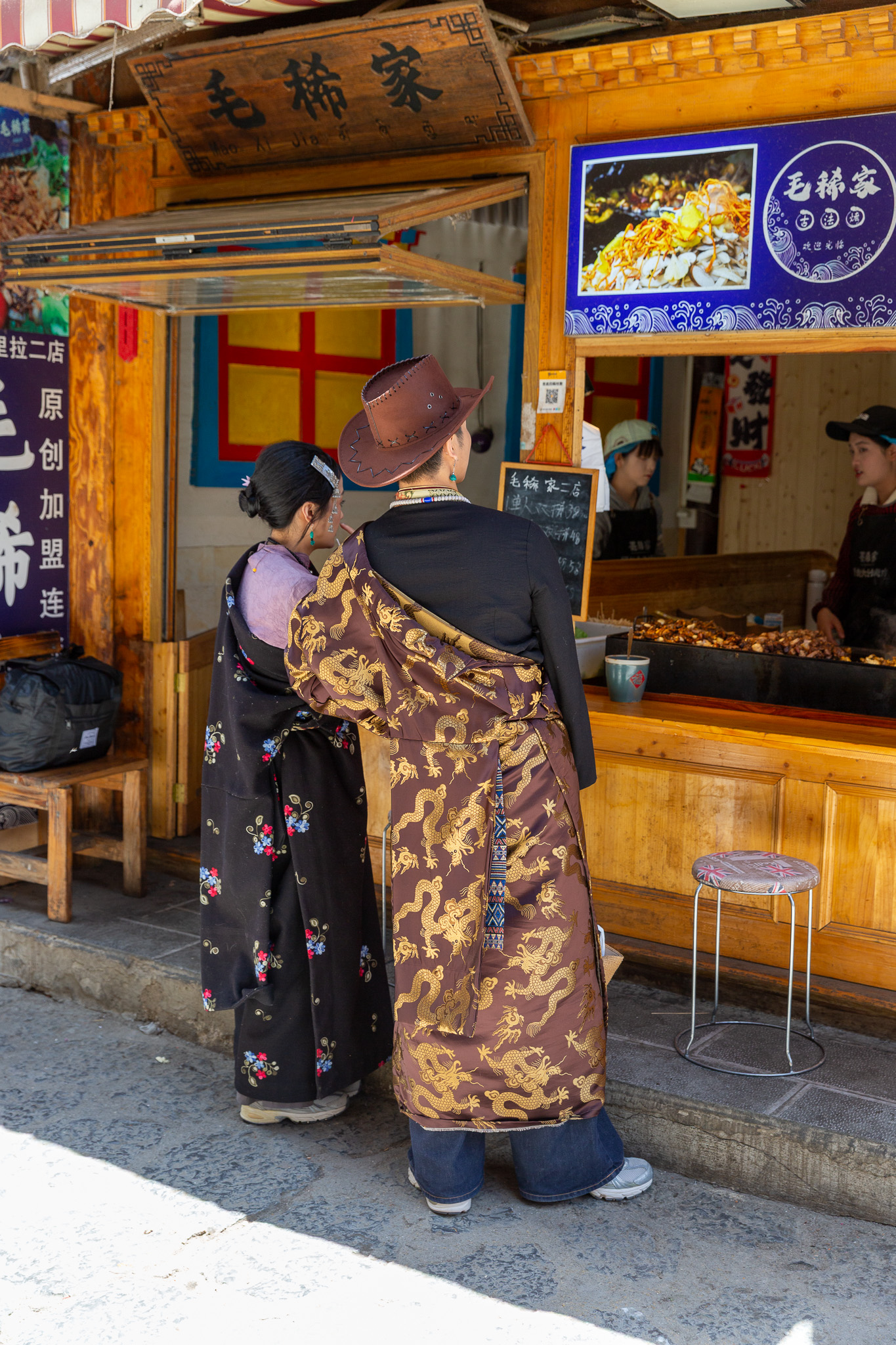 Pár v tradičnom tibetskom odeve a s kovbojským klobúkom stojí pri drevenom stánku s pouličným jedlom v meste Shangri-La.