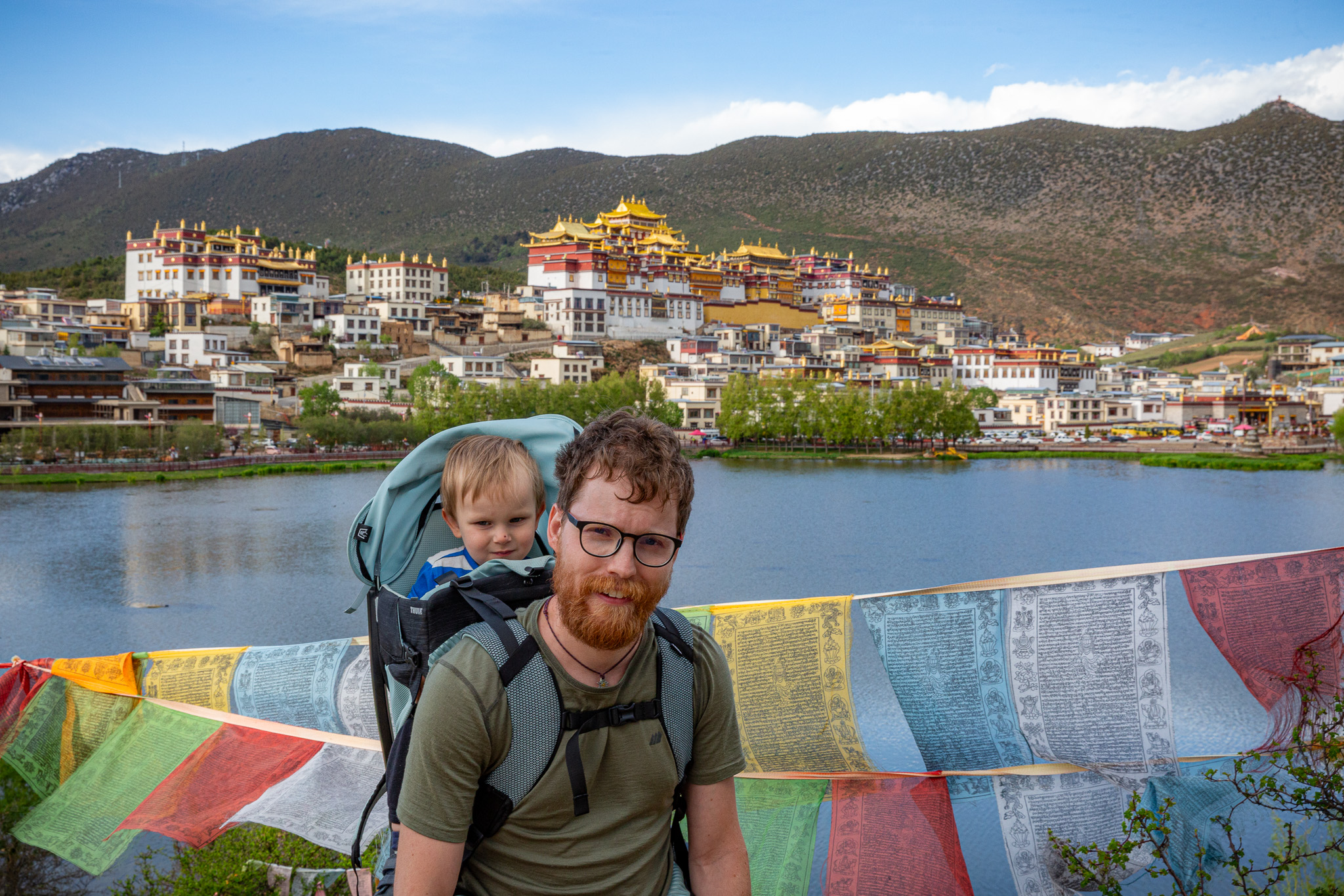 Muž s dieťaťom v nosiči stojí pred jazerom, za ktorým na kopci stojí celý areál Songzanlin Monastery, Shangri-La