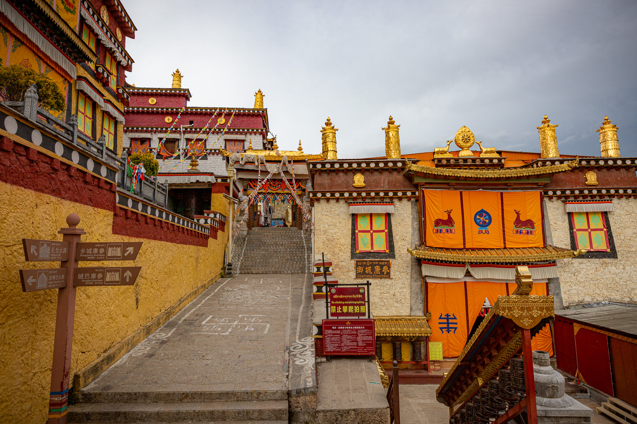 Kamenné schodisko vedúce do vnútra tibetského kláštora so žlto-červenými fasádami, zlatými prvkami na streche a smerovými tabuľami v popredí. Shangri-La