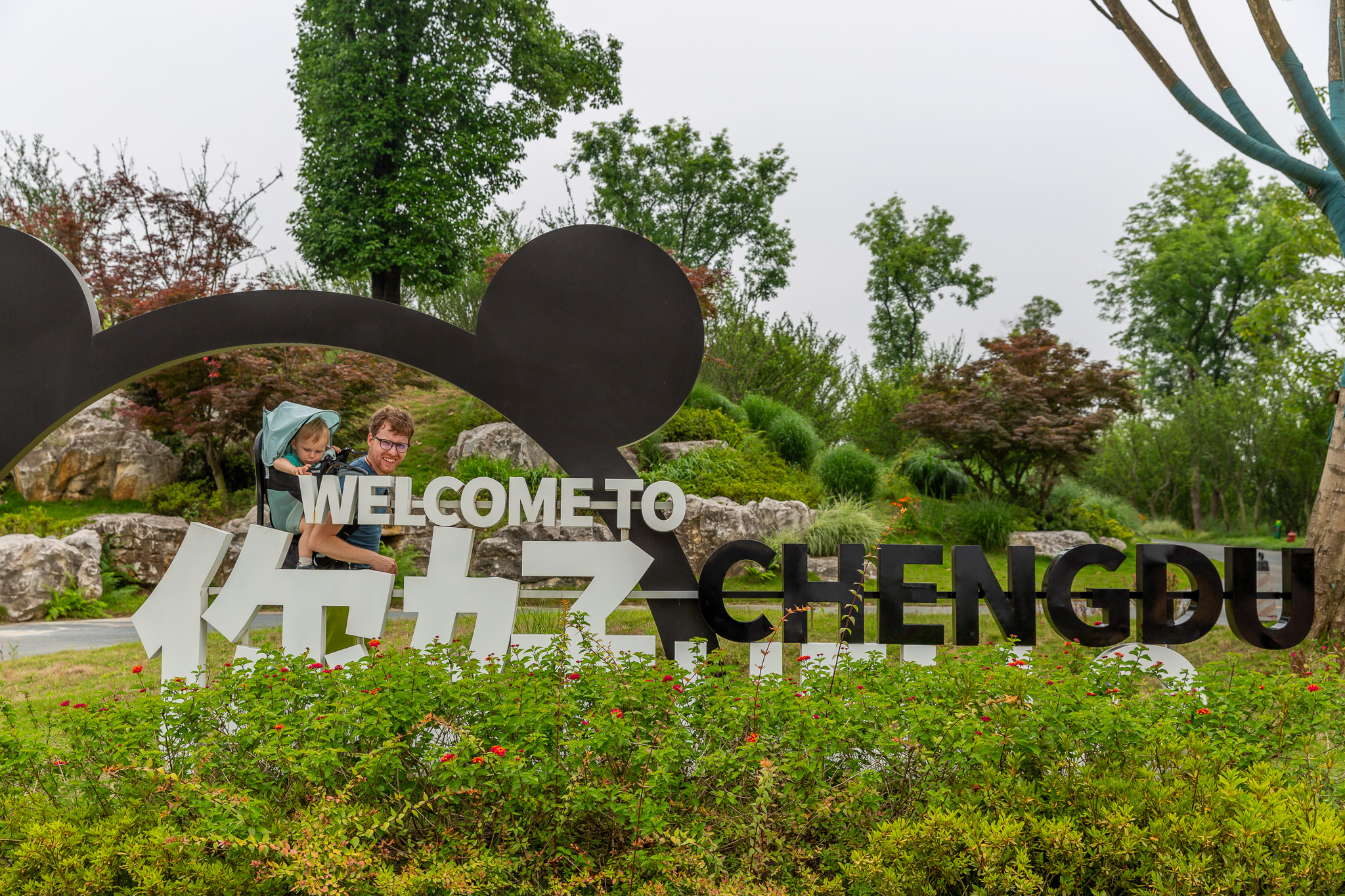 Muž s dieťaťom v nosiči stojí za nápisom Welcome to Chengdu. Okolo množstvo zelene. Chengdu Research Base of Giant Panda Breeding