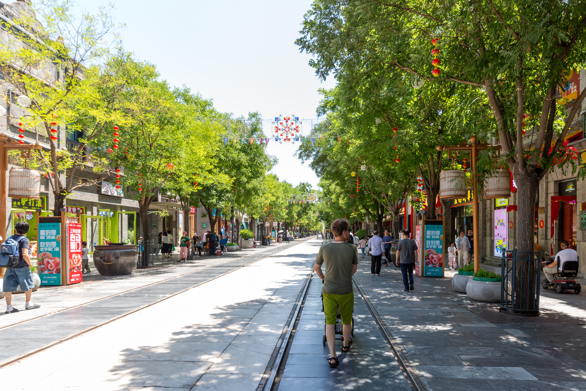 Muž tlačiaci kočík kráča vedľa električkovej koľaji po pešej zóne obklopenej stromami a obchodíkmi. Qianmen pedestrian Street
