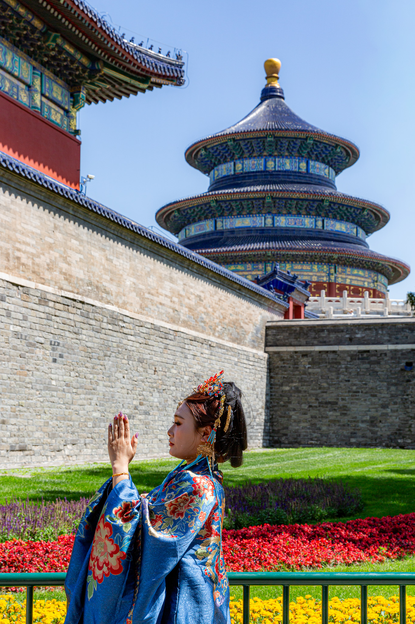Tmavovlasá žena so zložitým účesom odená do tradičného modrého kimona stojí s rukami zdvihnutými oproti sebe pred Temple of Heaven. Peking